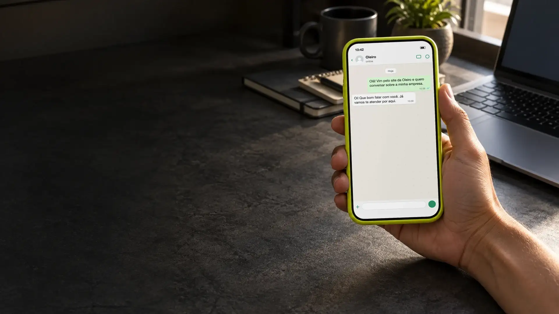 Mão segurando o celular com uma conversa de WhatsApp aberta, ao lado do notebook na mesa do escritório