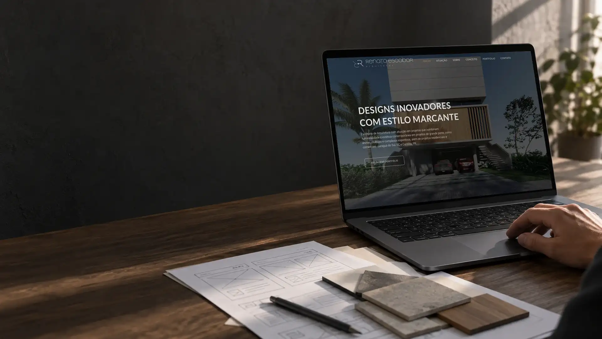 Mão no notebook com o site de um escritório de arquitetura aberto, sobre rascunhos do layout e amostras de material